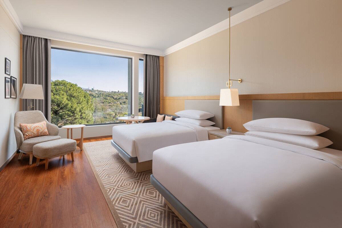 Habitación de hotel con dos camas, una silla, una mesita y vistas a zonas verdes a través de amplias ventanas.
