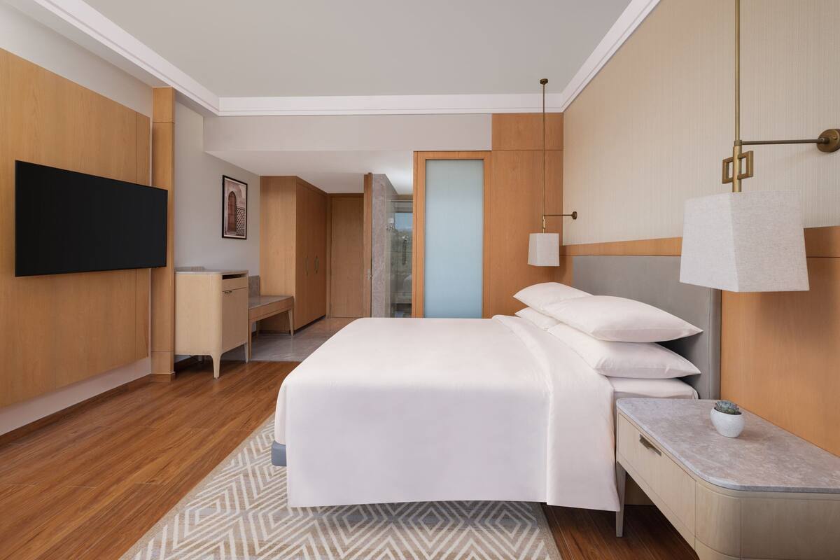 Una habitación de hotel moderna con una cama grande, un escritorio, un televisor de pantalla plana montado en la pared y un baño visible a través de una puerta de cristal.