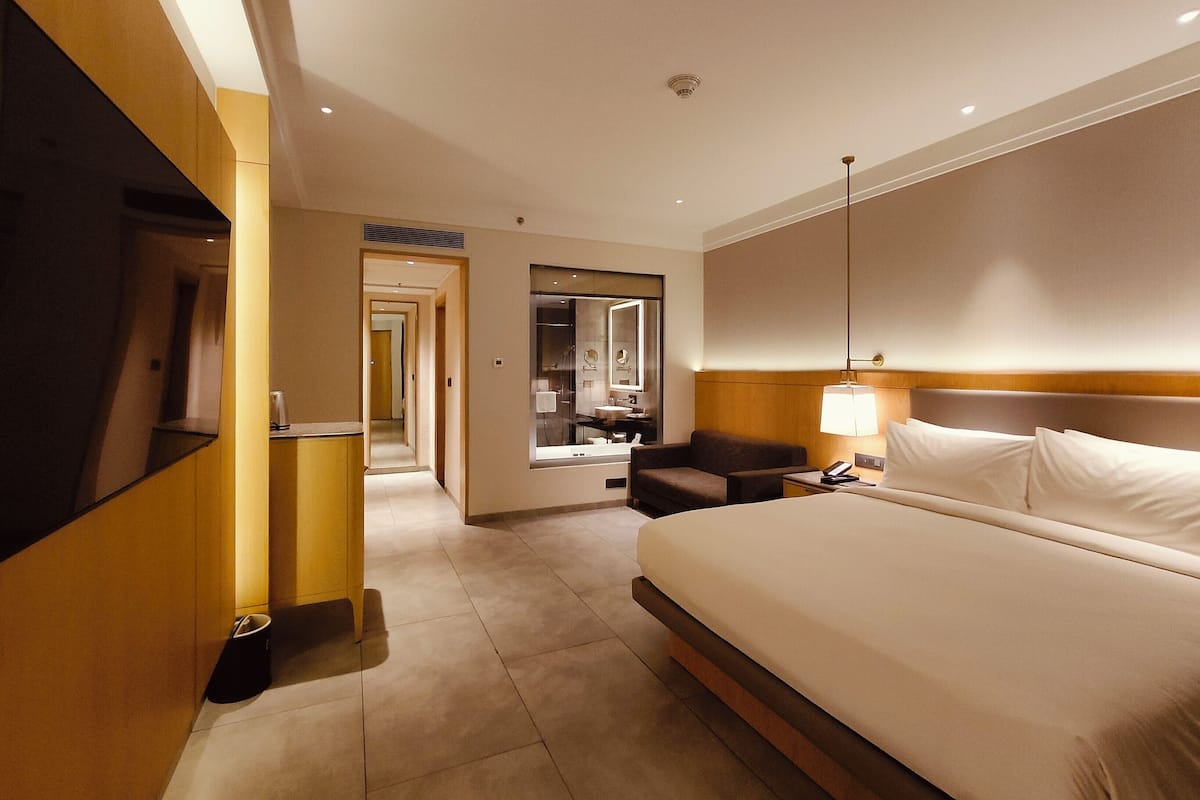 Habitación de hotel con una cama grande, un escritorio con lámpara, un espejo y vistas al pasillo.