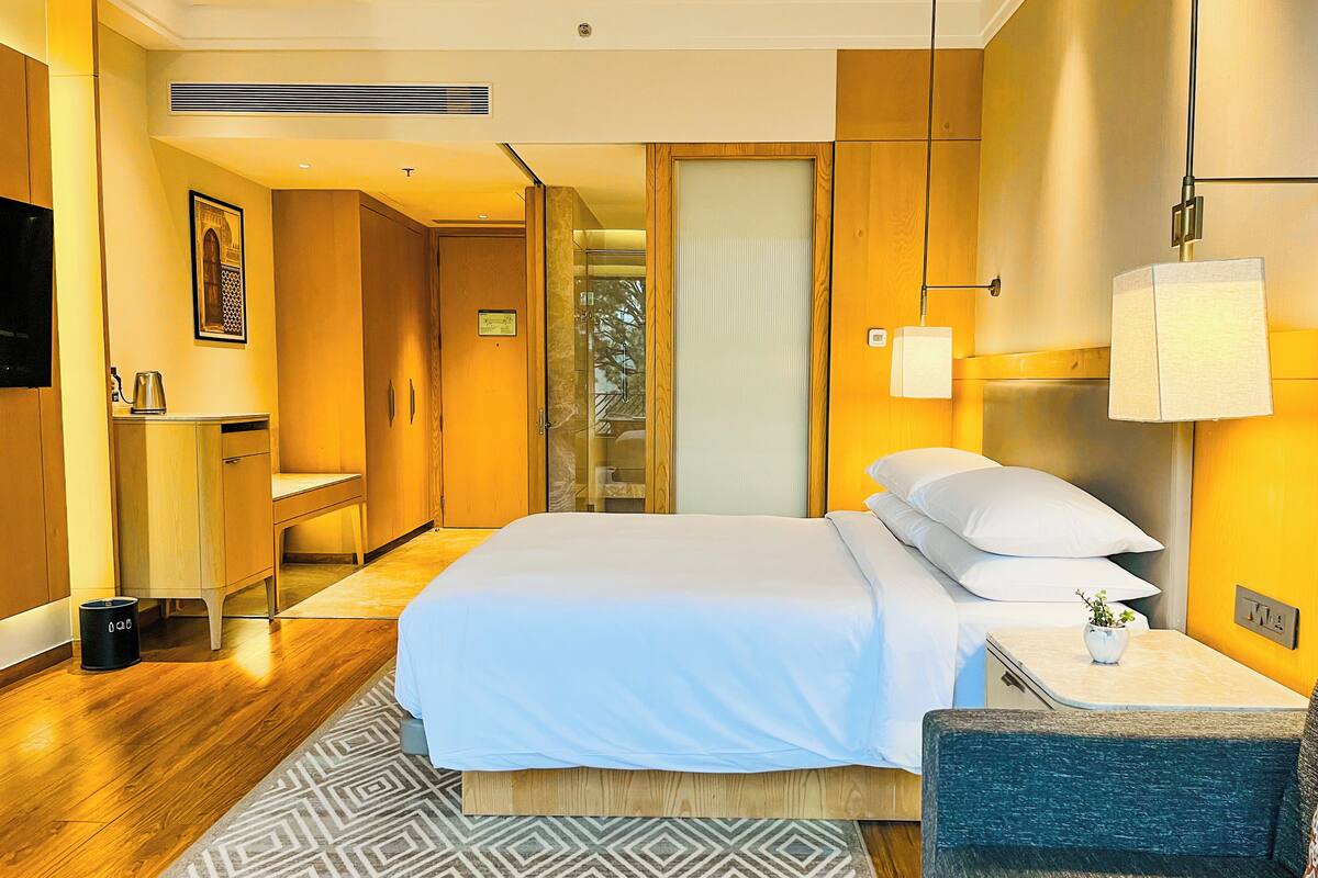 Una habitación de hotel moderna con una cama grande, un escritorio y una silla. Hay un televisor en la pared y una ventana con vistas a árboles.