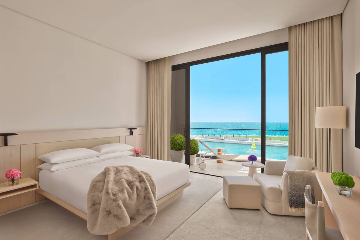 A modern hotel room with a large bed, a sitting area with a view of the ocean, and a balcony with a pool.