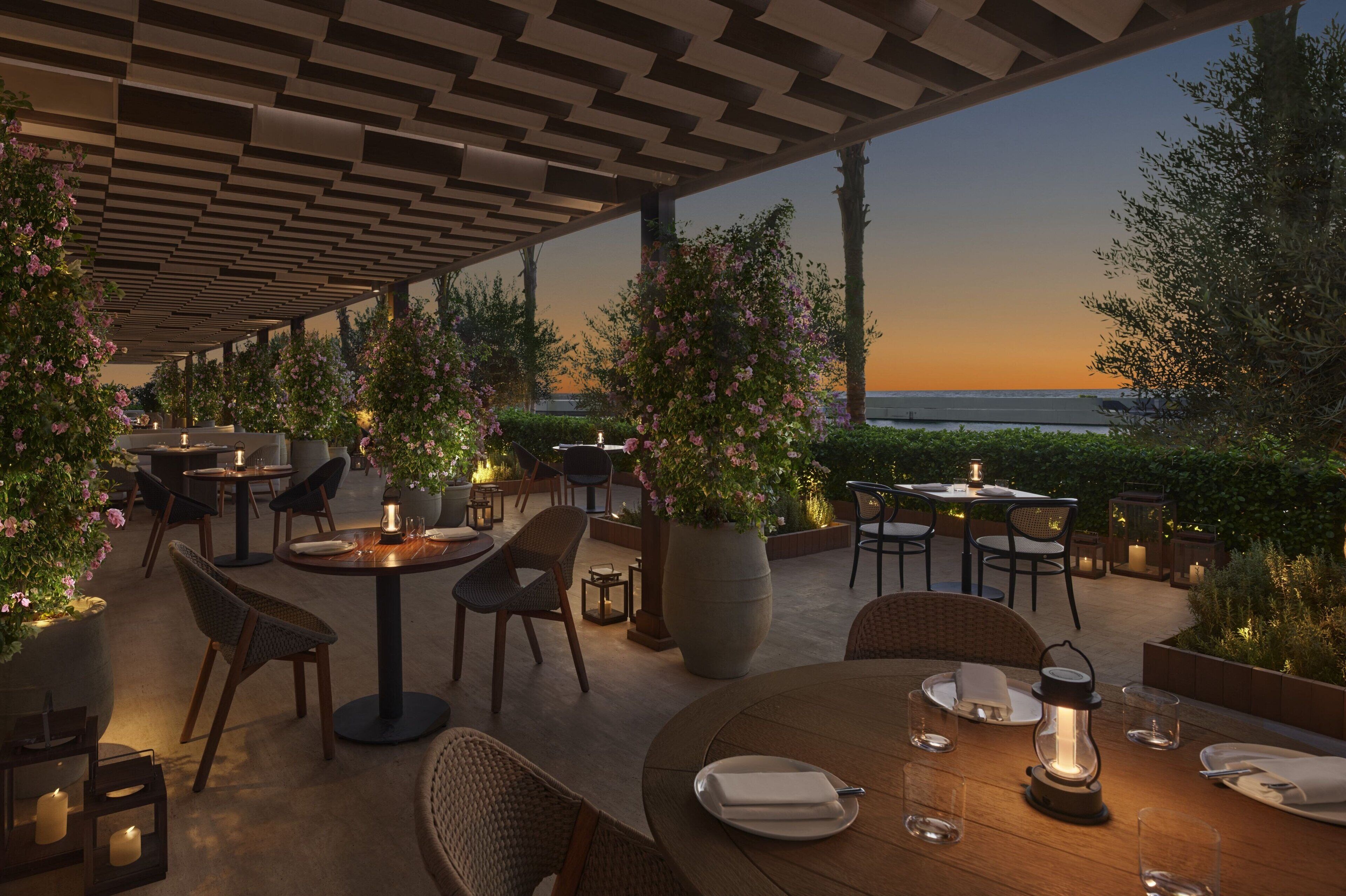 Un espacio de comedor al aire libre con mesas y sillas, rodeado de vegetación y vistas al mar al atardecer.