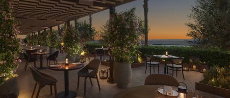 Un espacio de comedor al aire libre con mesas y sillas, rodeado de vegetación y vistas al mar al atardecer.