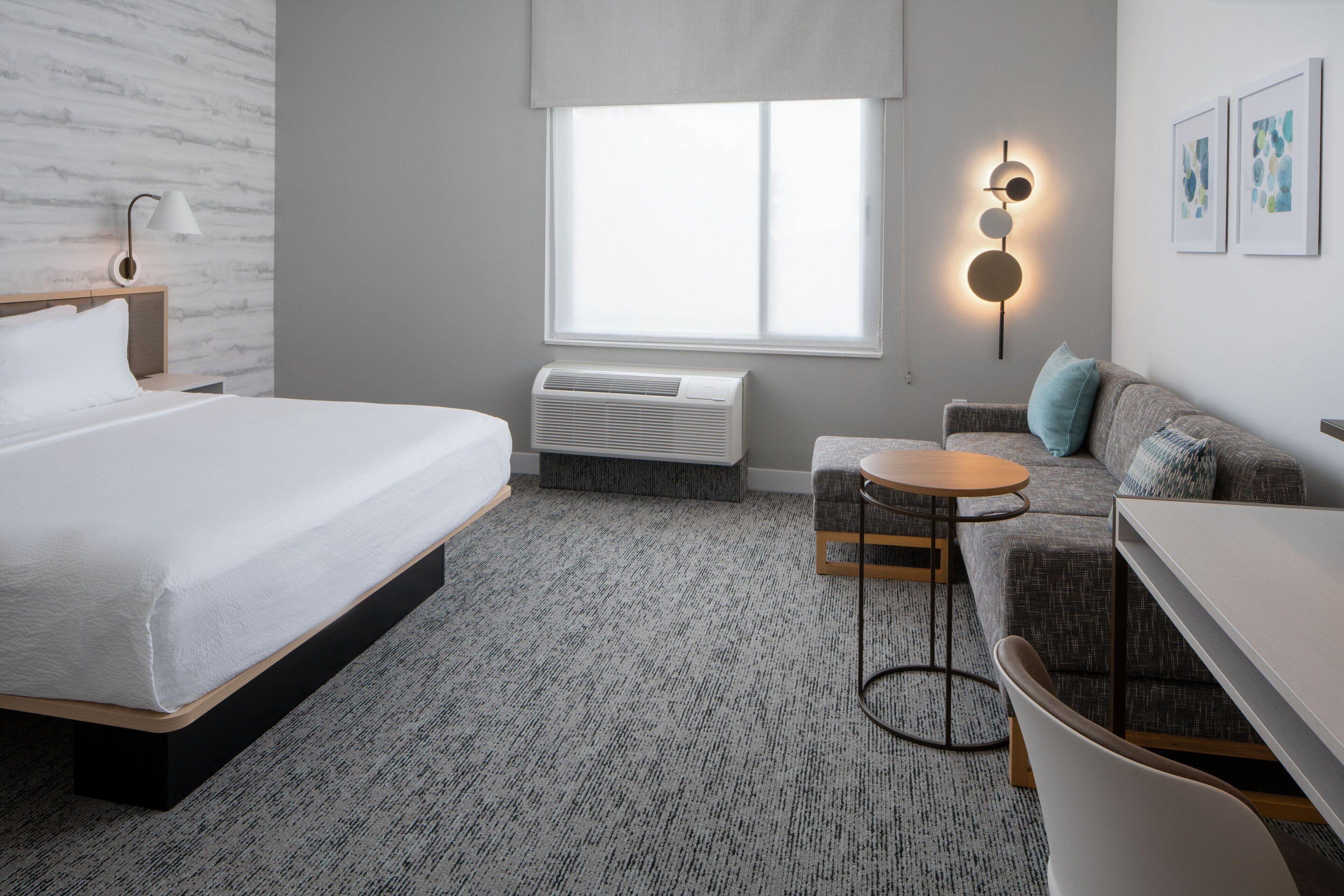 A modern hotel room with a bed, a sofa, a small table, and a wall-mounted lamp.