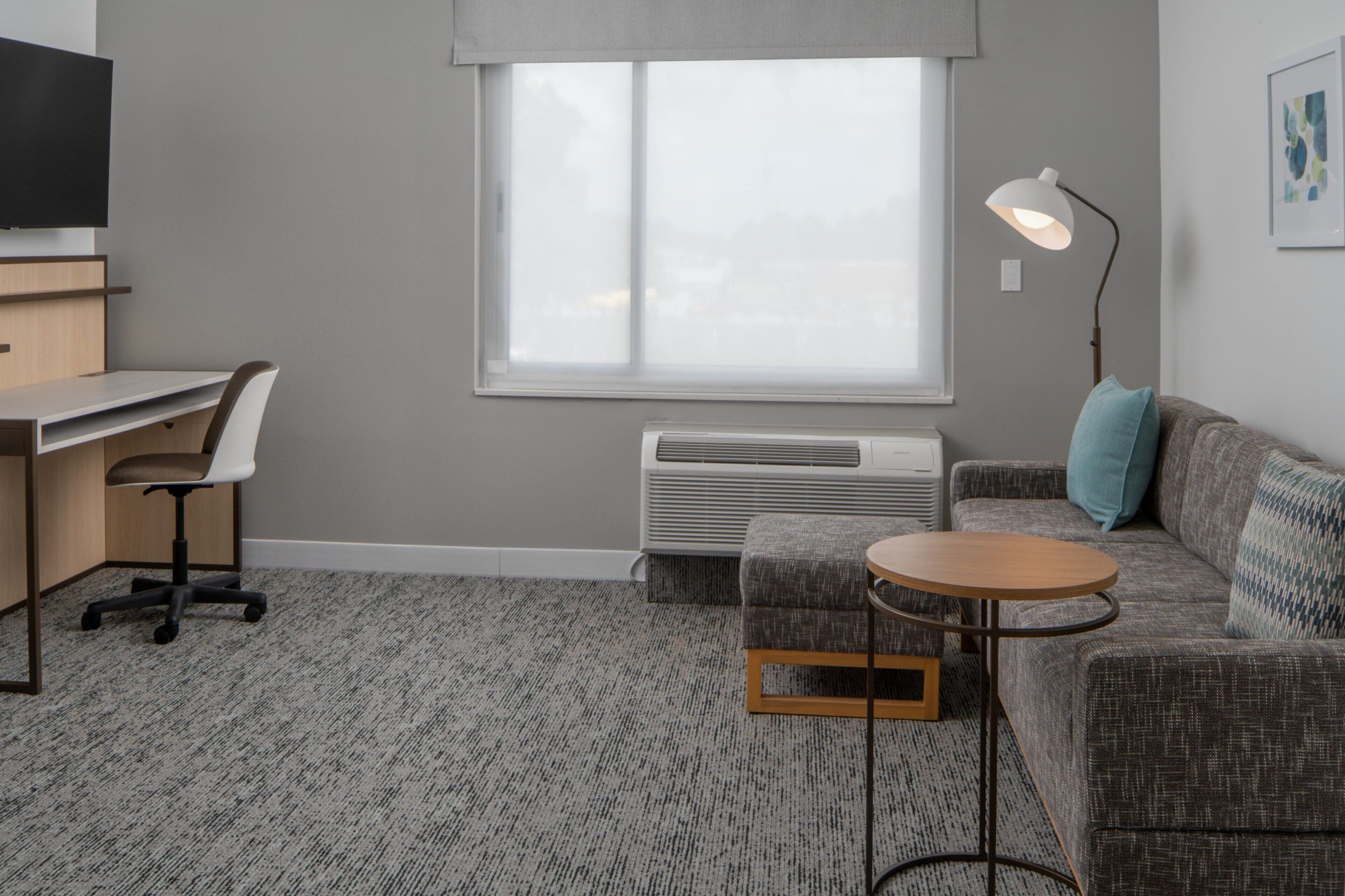 A modern living room with a grey carpet, a grey sofa, a round wooden table, a grey armchair, a flat-screen TV mounted on the wall, and a window with a view of the sky.