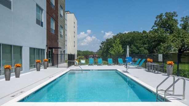 Une piscine rectangulaire à l’eau d’un bleu limpide, entourée d’une terrasse blanche. On y trouve plusieurs chaises longues et tables, et une clôture délimite l’espace.