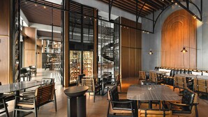 A spacious restaurant with high ceilings, wooden beams, and a central spiral staircase. Tables and chairs are arranged for dining, and a wine cooler is visible in the background.