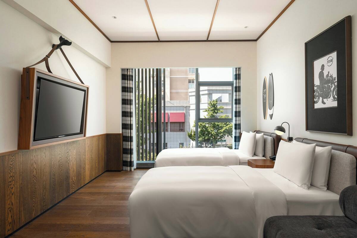 A hotel room with two beds, a TV mounted on the wall, a large window with curtains, and a framed picture on the wall.