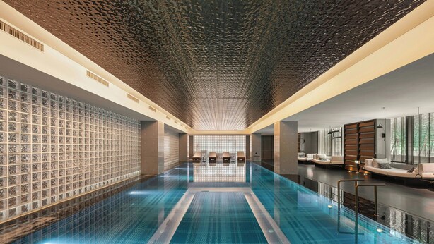 A modern indoor swimming pool with a geometric patterned floor, a textured ceiling, and a seating area with a sofa and chairs.