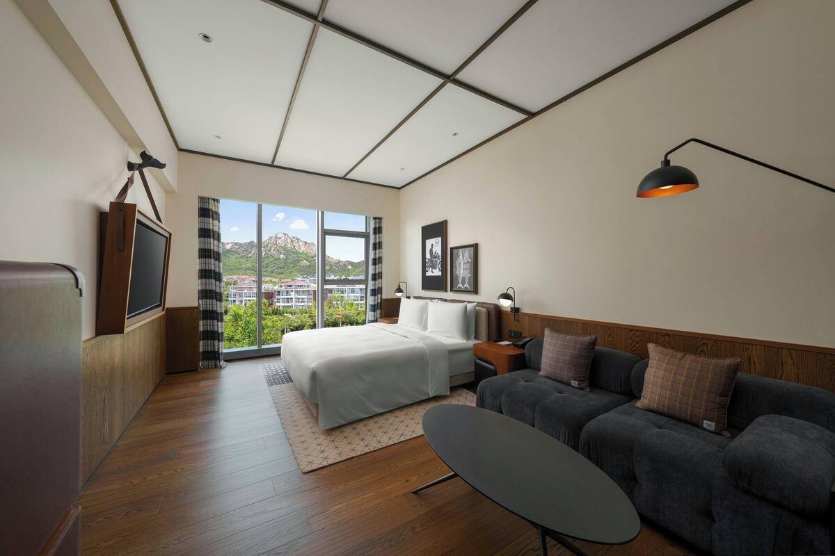 A modern hotel room with a large bed, a sofa, a small table, and a view of the outdoors.