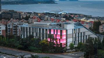 Un edificio moderno con una fachada iluminada en rosa, rodeado de edificaciones residenciales y vistas al mar.