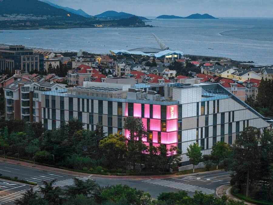 Un edificio moderno con una fachada iluminada en rosa, rodeado de edificaciones residenciales y vistas al mar.