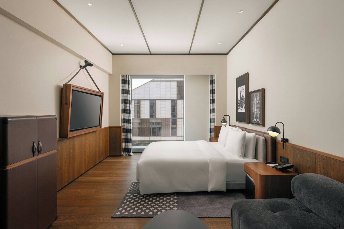 A modern hotel room with a large bed, a TV mounted on the wall, a desk with a lamp, and a view of the city through the window.