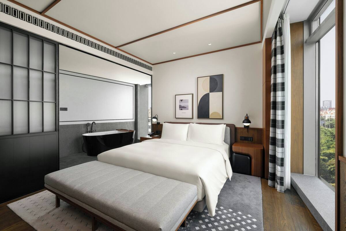 A modern hotel room with a large bed, bedside tables, a desk, and a view of the city.