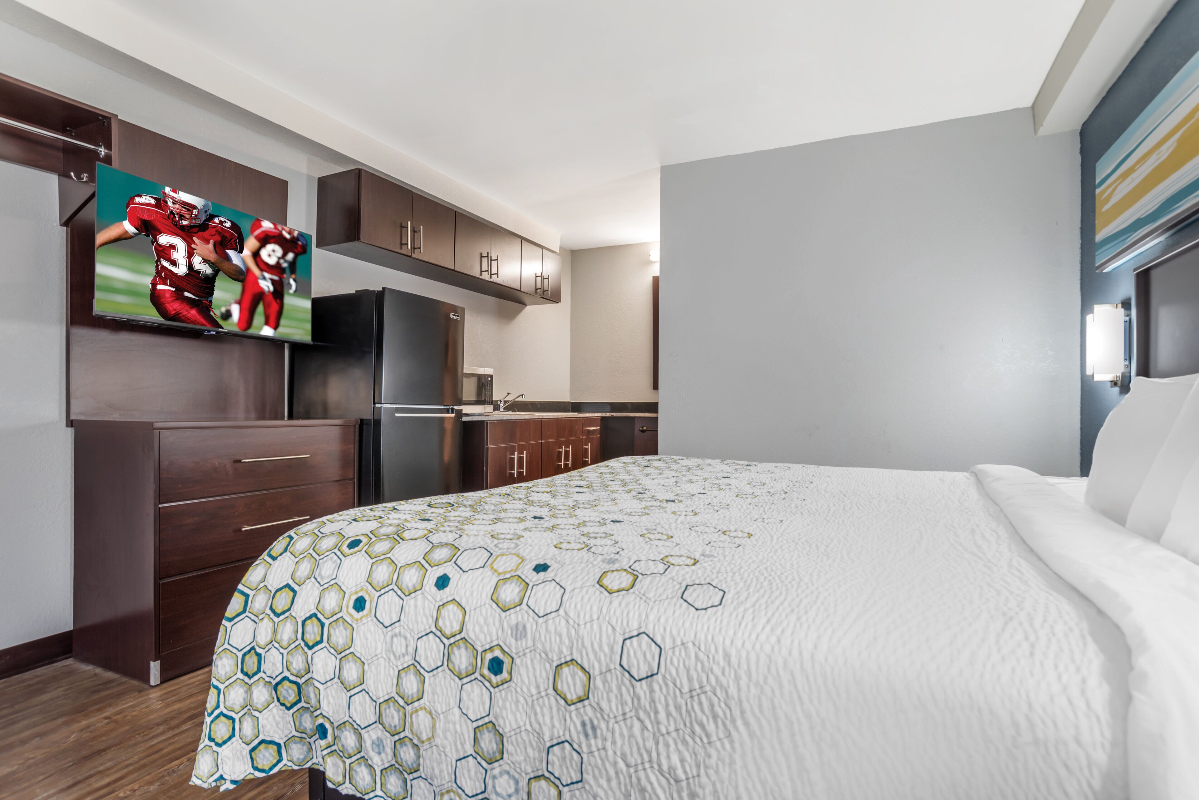 A hotel room with a bed, a TV mounted on the wall, a refrigerator, and wooden cabinets.