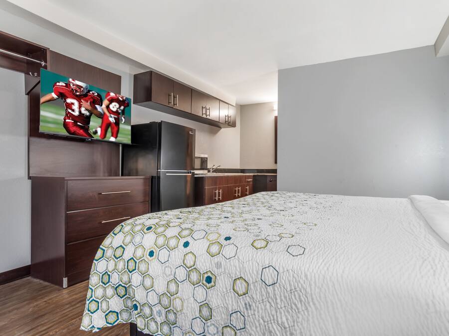 A hotel room with a bed, a TV mounted on the wall, a refrigerator, and wooden cabinets.