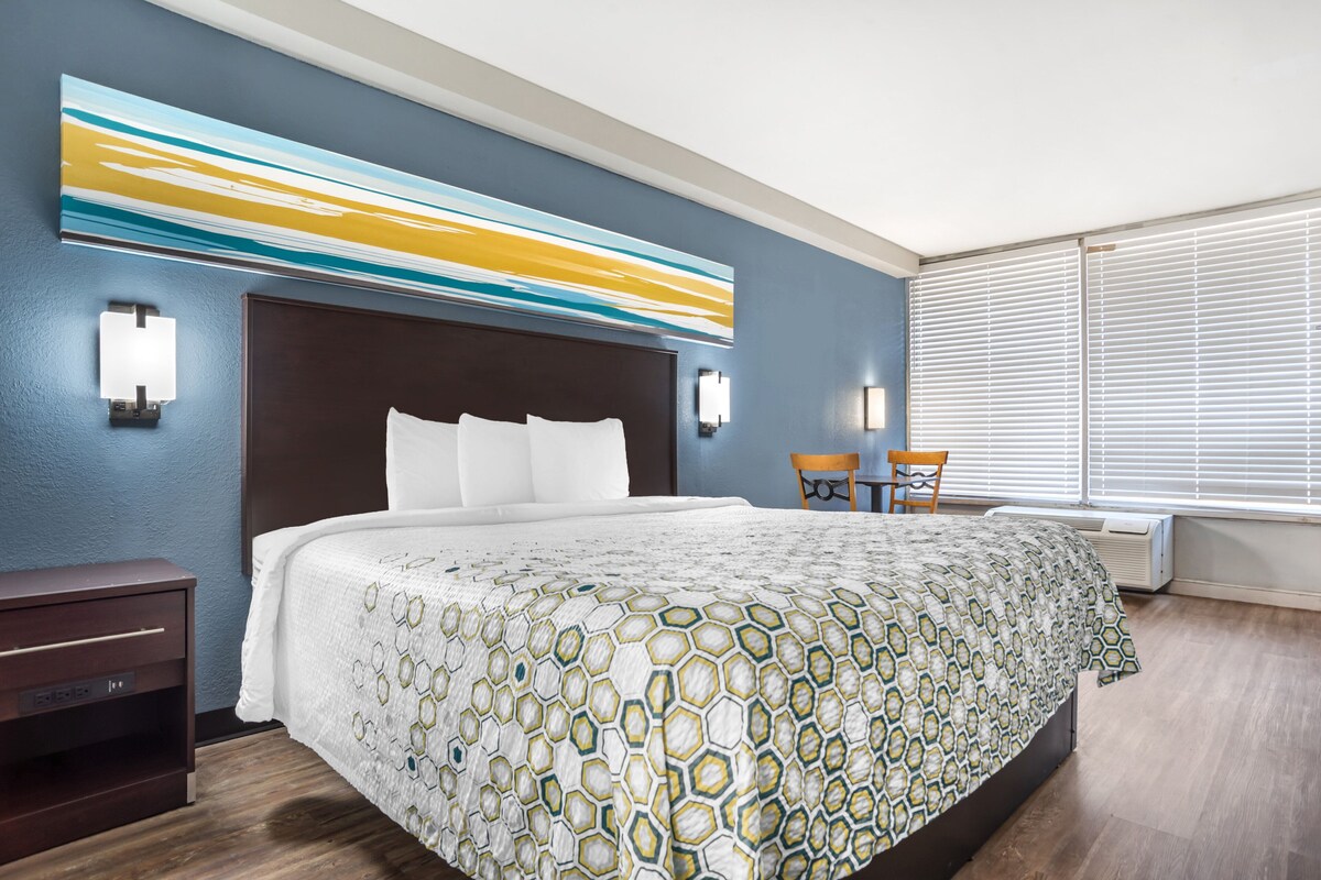 A hotel room with a large bed, a nightstand, a chair, and a wall-mounted artwork.