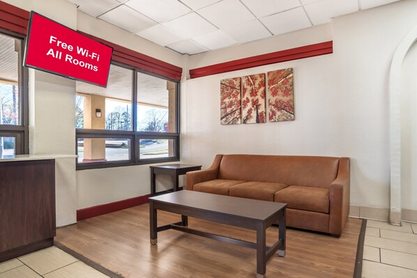 A modern living room with a brown sofa, a wooden coffee table, and a red sign indicating free Wi-Fi availability. - HomeTowne Studios by Red Roof Montgomery - Midtown (Montgomery)