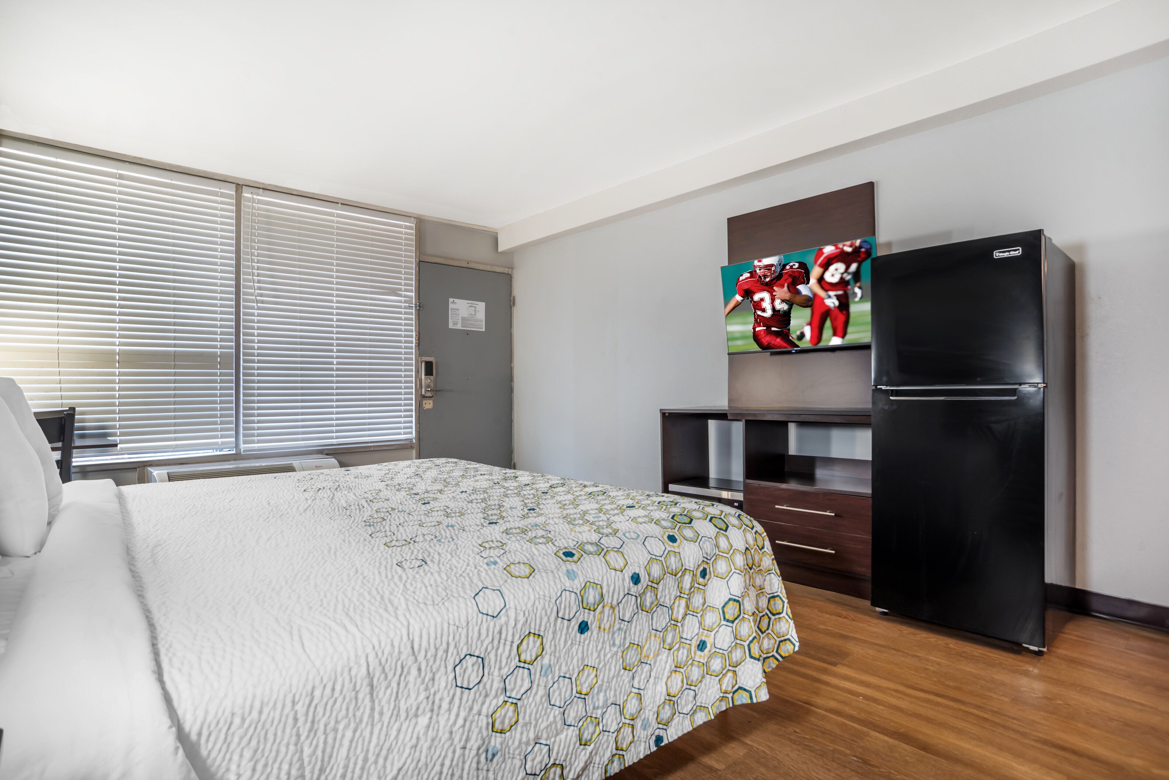 A hotel room with a bed, a refrigerator, a television, and a window with blinds.