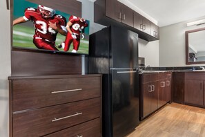 A modern kitchen with dark wood cabinetry, a stainless steel refrigerator, and a flat-screen TV mounted above the cabinetry. - HomeTowne Studios by Red Roof Montgomery - Midtown (Montgomery)
