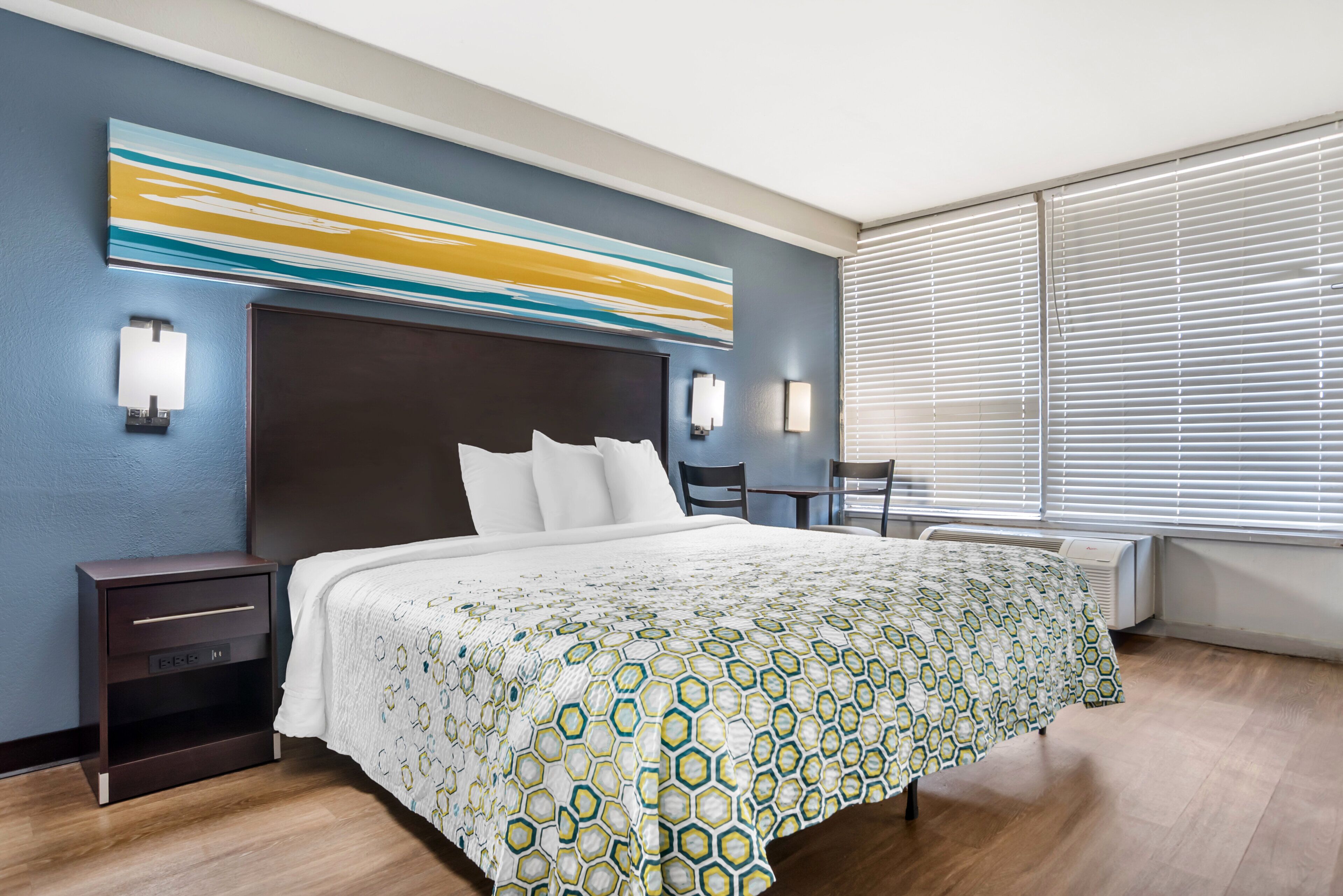 Ein Hotelzimmer mit einem großen Bett, einem Kopfteil, Nachttischen, einem Schreibtisch und einem Fenster mit Jalousien.