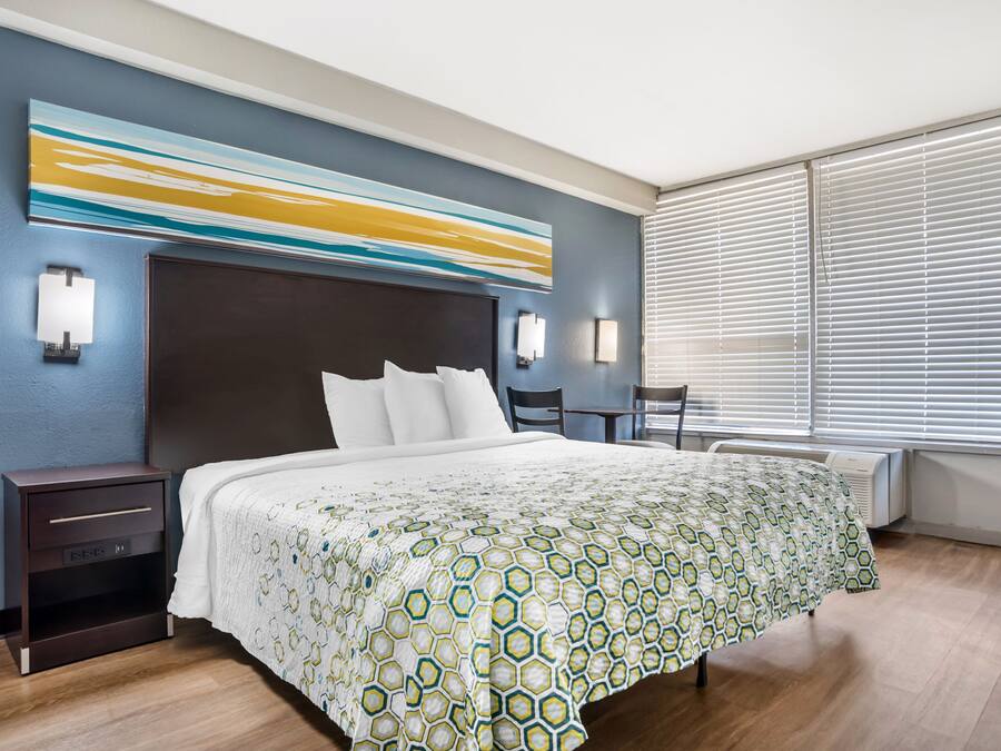 Ein Hotelzimmer mit einem großen Bett, einem Kopfteil, Nachttischen, einem Schreibtisch und einem Fenster mit Jalousien.