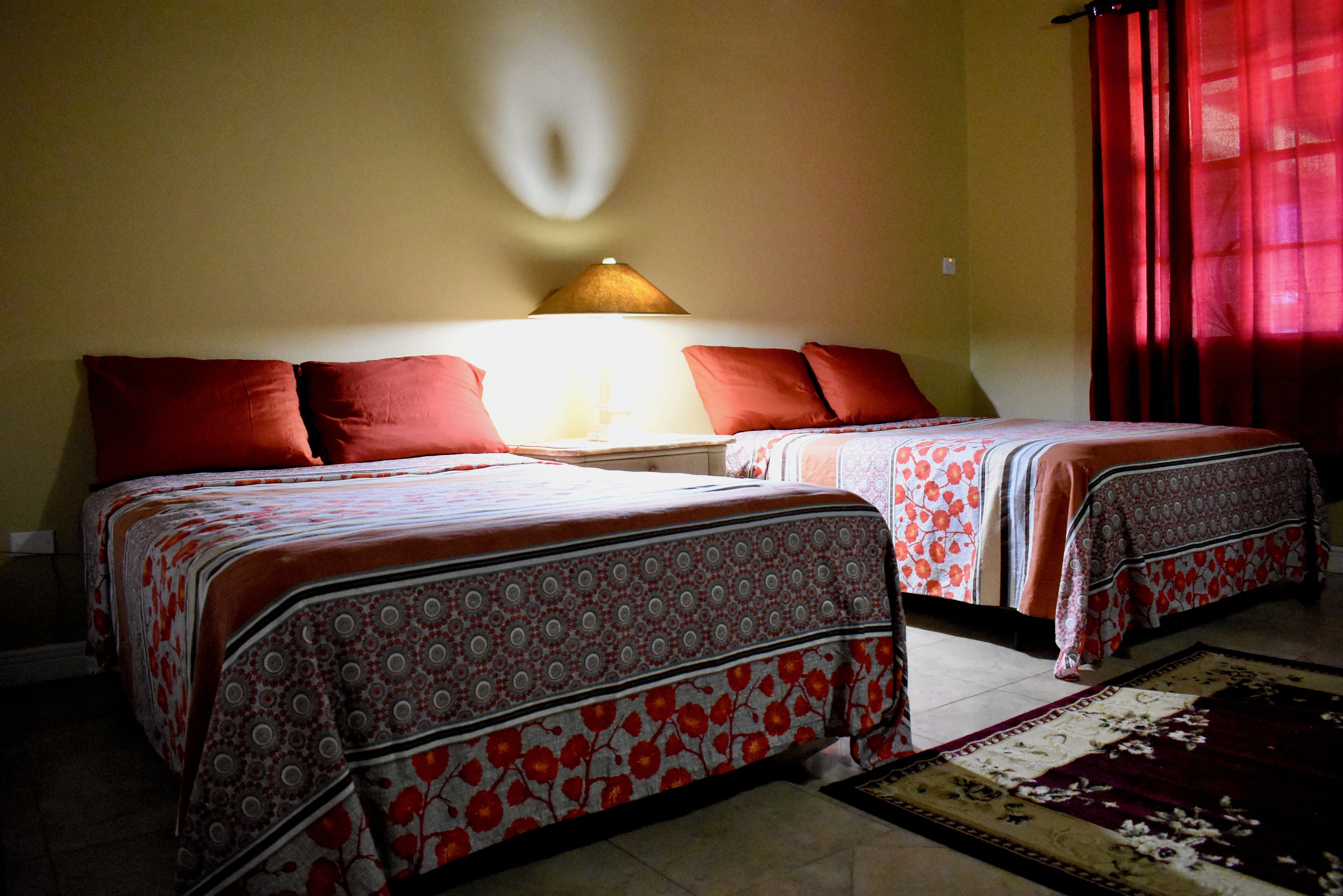 A hotel room with two beds, each with a patterned bedspread and red pillows. There is a lamp on the nightstand between the beds, and a red curtain on the window.