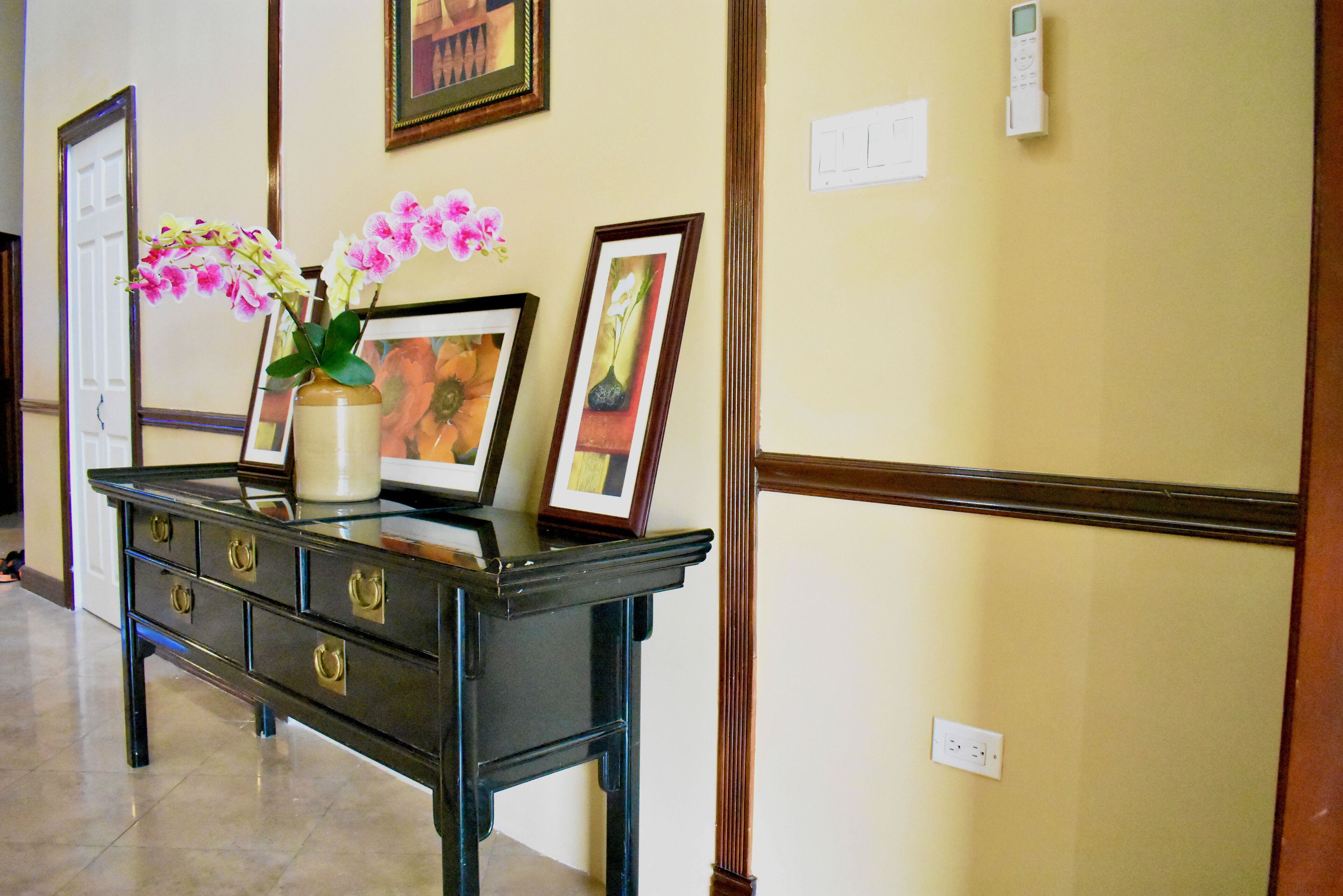 Une console en bois noir, sur laquelle sont posés un vase rempli d’orchidées roses, deux tableaux encadrés et un vase blanc.