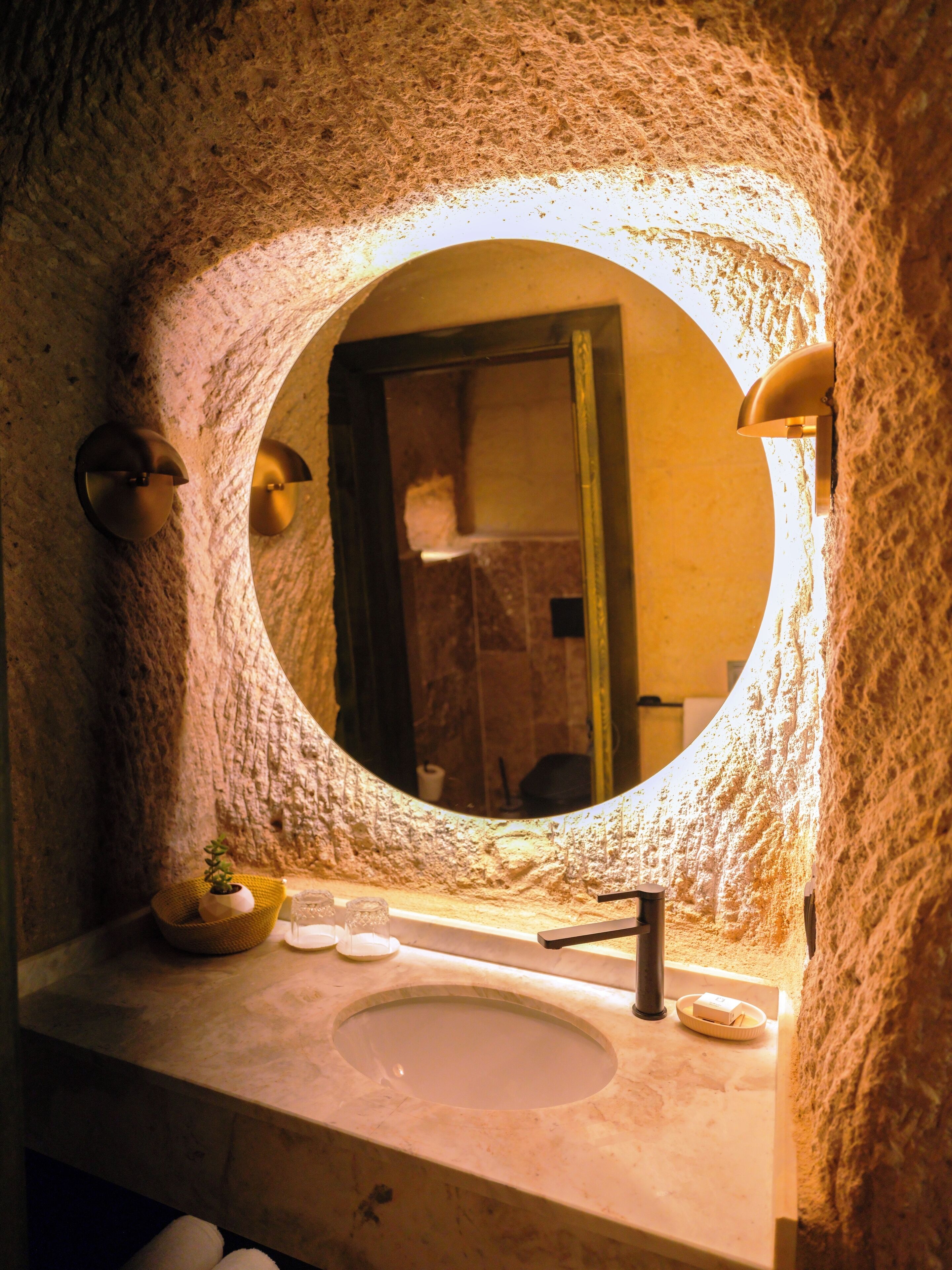 A bathroom with a stone wall, a round mirror, a sink, and a small plant.