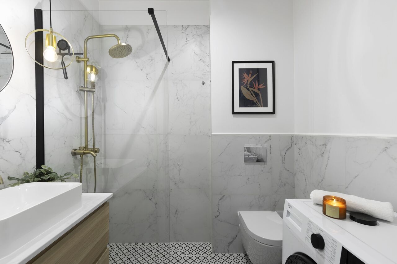Un baño moderno con pared de mármol, un lavabo blanco, ducha con detalles dorados y un cuadro enmarcado en la pared.
