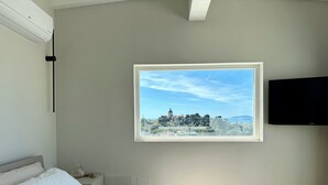 Quarto com cama, televisão, janela com vista para a cidade e ventoinha no teto.