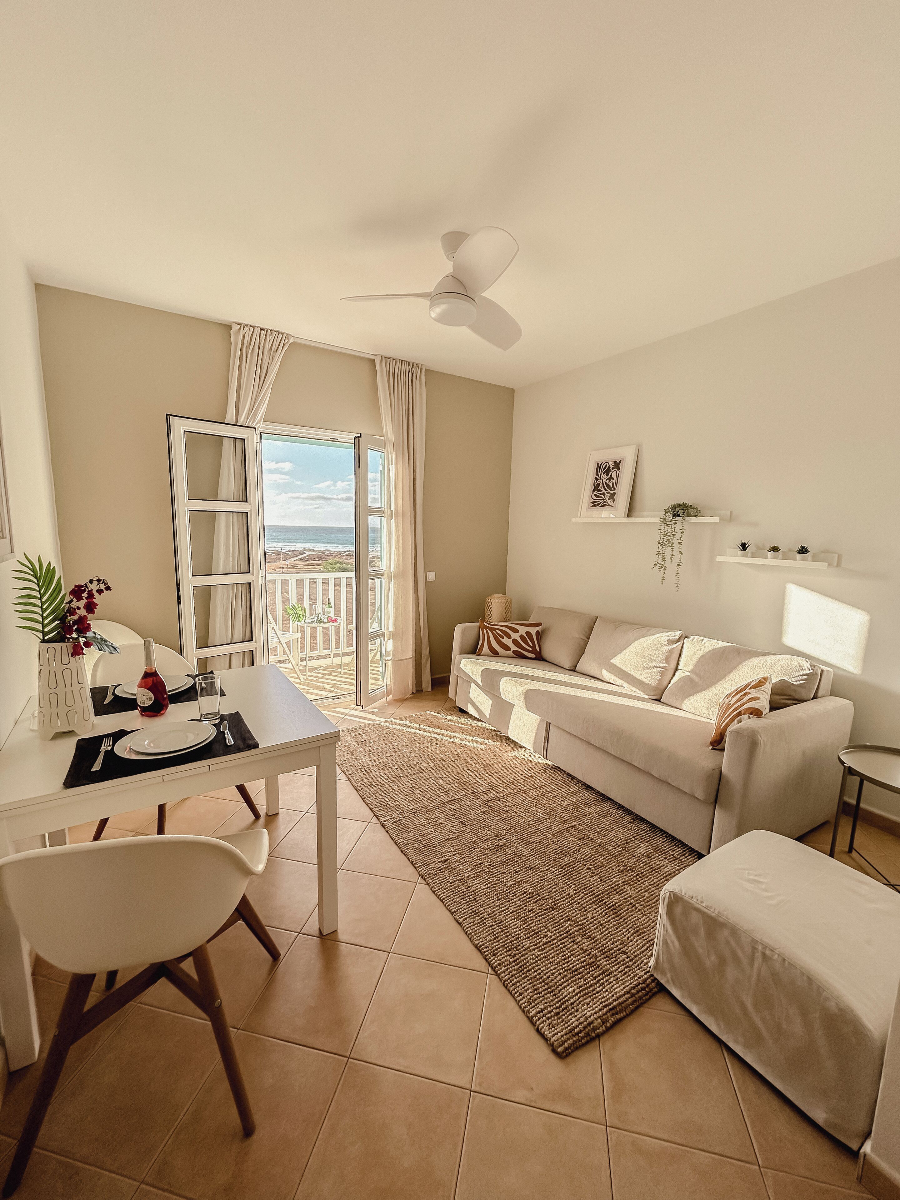 Una sala de estar moderna con comedor, un sofá blanco, un ventilador de techo y vista a la playa a través del ventanal.