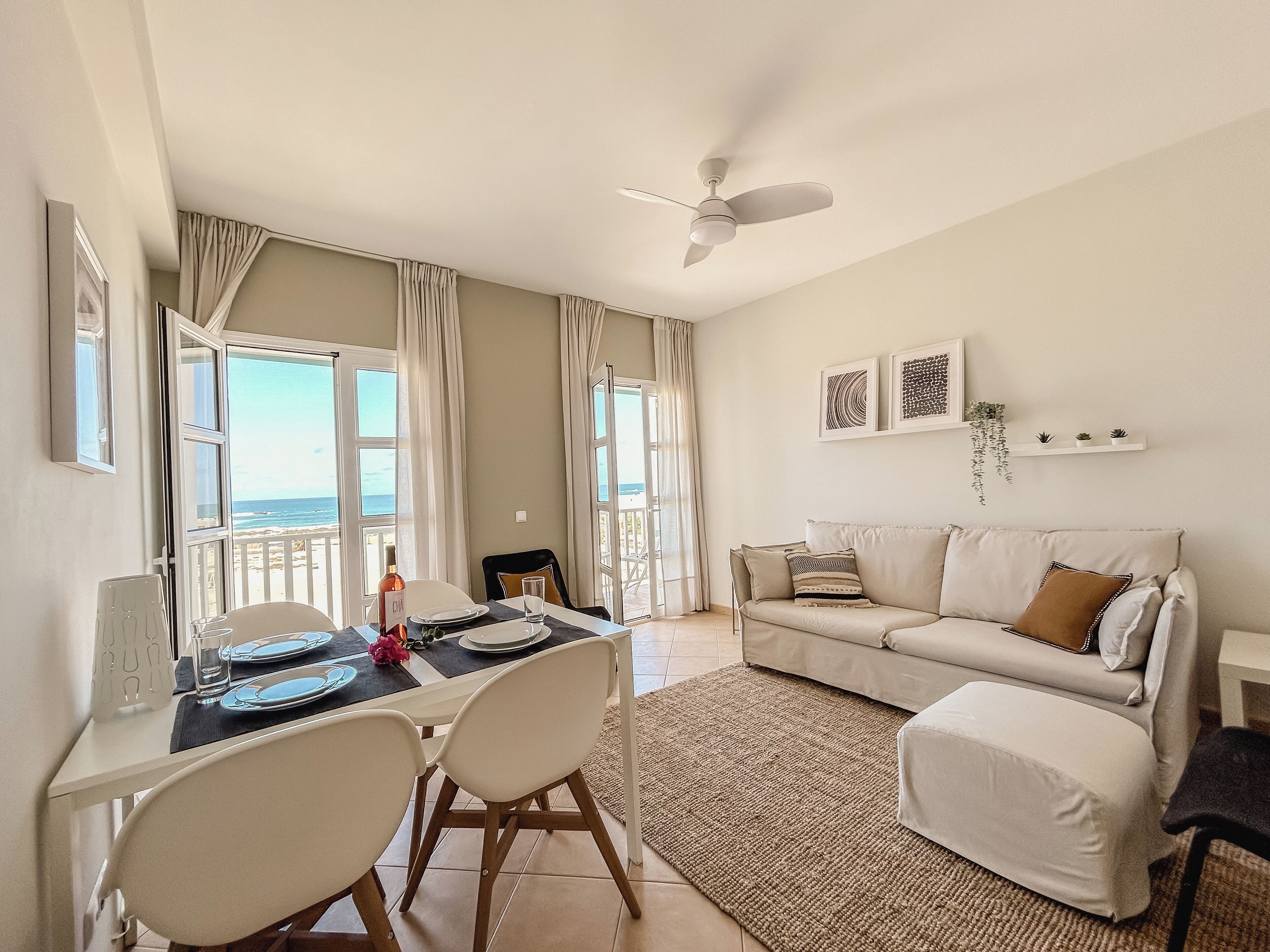 A dining area with a table set for two, a ceiling fan, and a view of the ocean through large windows.