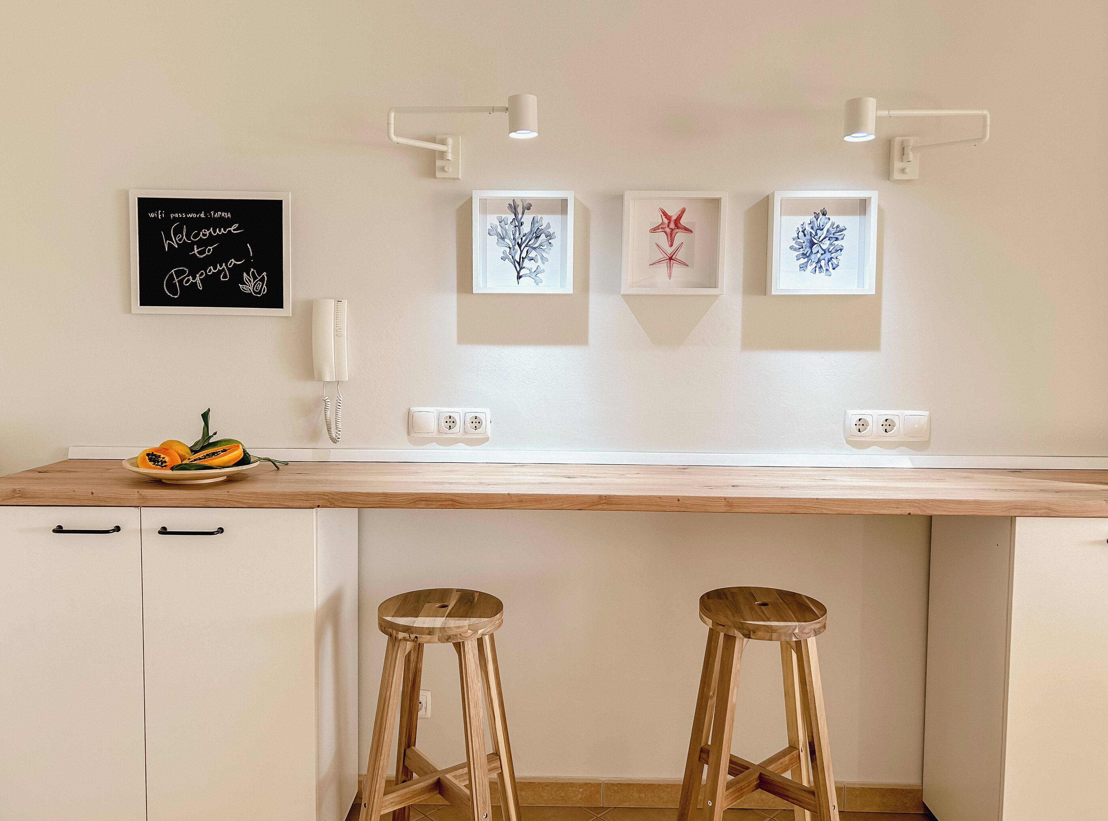 A kitchen with a wooden countertop, two bar stools, a fruit bowl, a wall-mounted phone, and framed artwork.