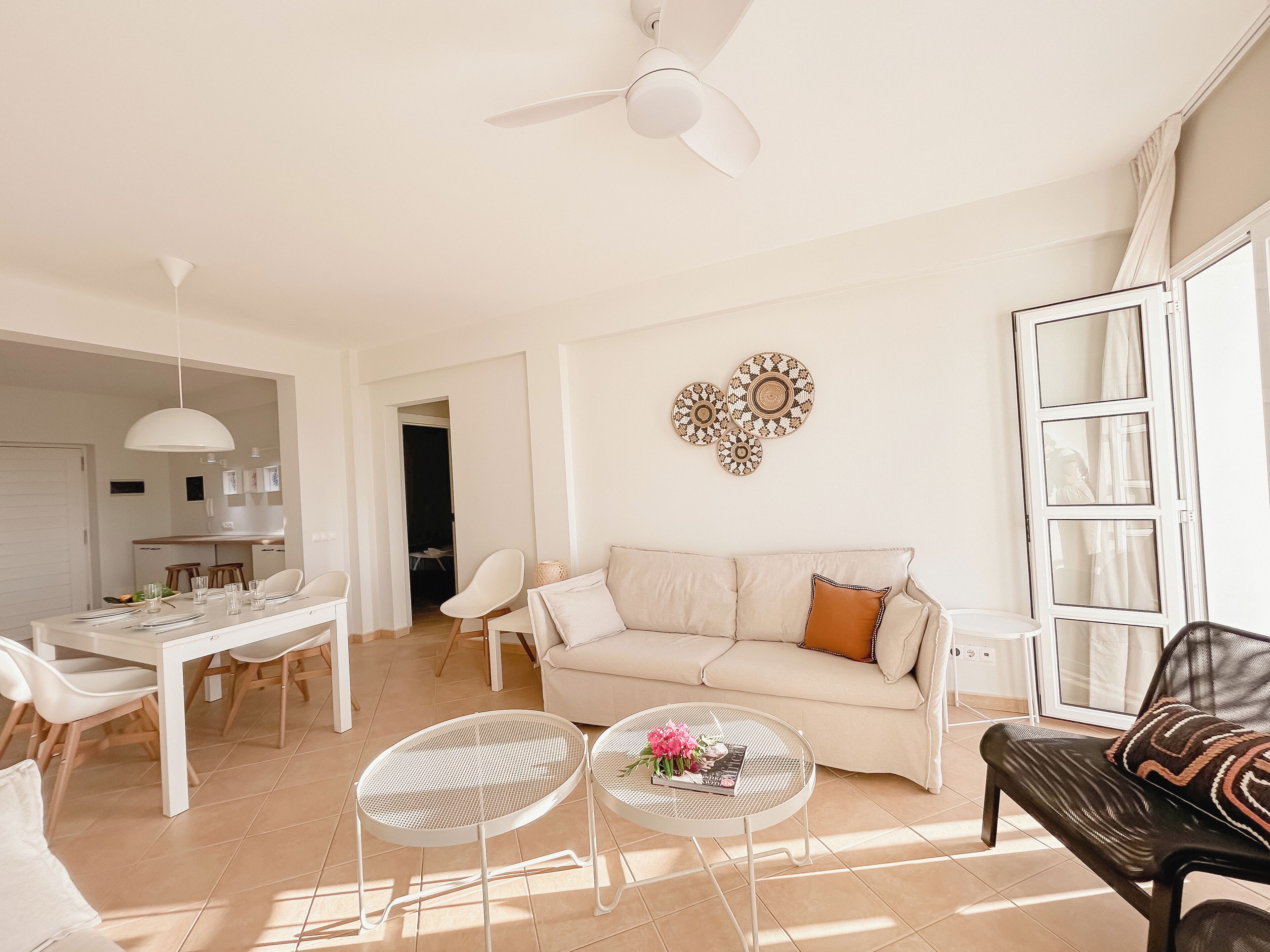 Una sala de estar luminosa con un sofá, dos mesitas, un comedor con mesa y sillas, y un ventilador de techo.