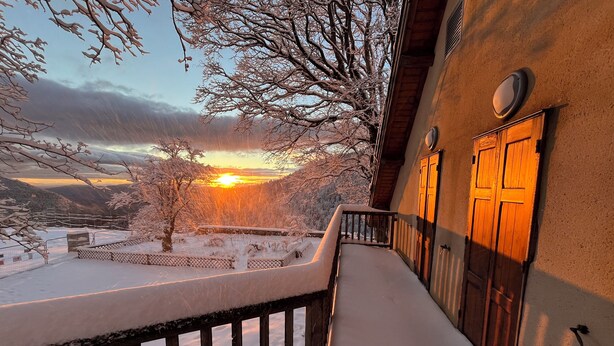 Een balkon met sneeuw bedekte relingen, een houten deur en uitzicht op een besneeerd landschap bij zonsondergang.