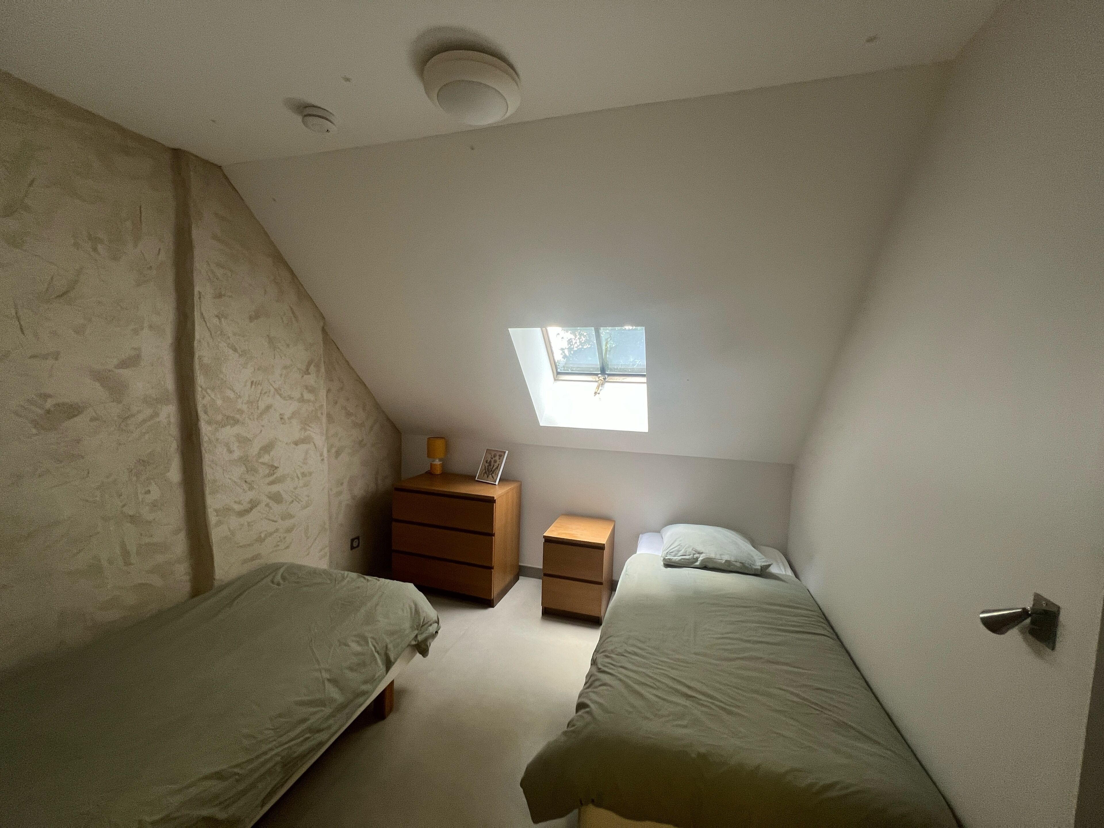 A small, dimly lit room with two single beds, a small wooden nightstand, a desk, and a window with a view of trees.