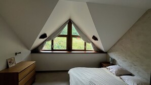 Een slaapkamer met een schuine kap, een raam met gordijnen, een houten dressoir, een nachtkastje en een bed met kussens.