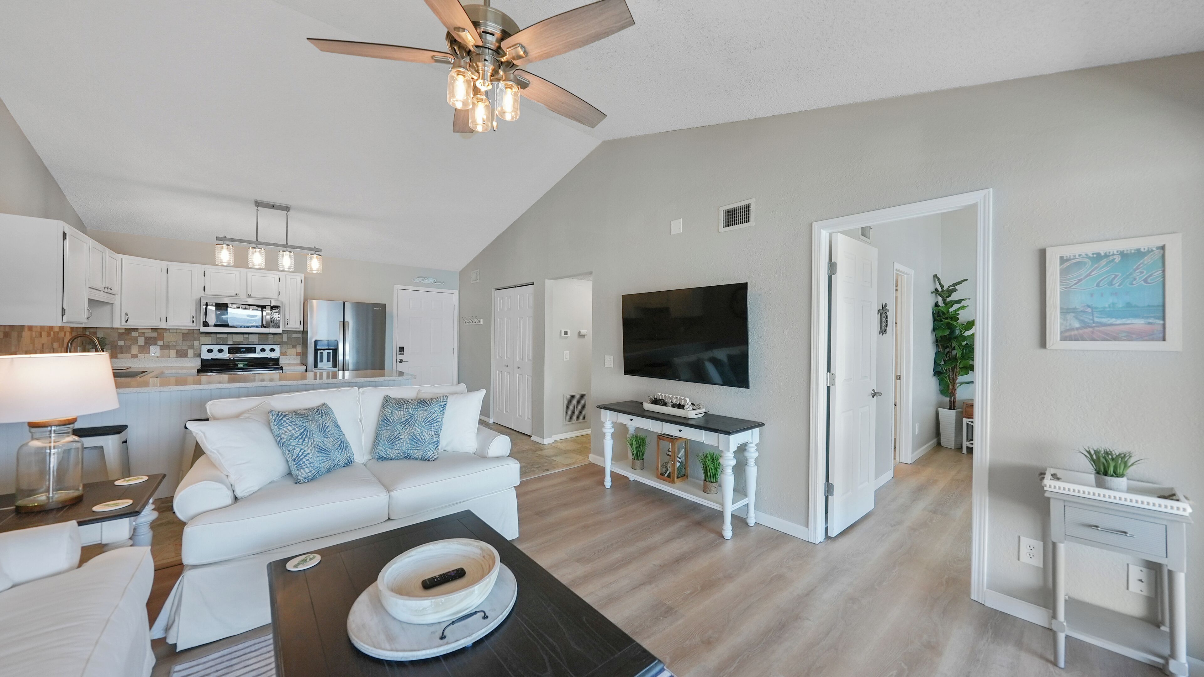 A spacious living room with a white sofa, a coffee table, and a TV. There is a kitchen area with white cabinets and a dining table.
