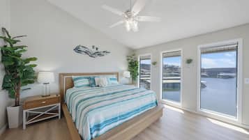 A spacious bedroom with a large bed, a ceiling fan, and a view of a body of water through the windows.