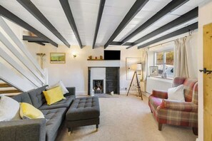 A living room with a fireplace, a sofa, an ottoman, and a plaid armchair. - Cosy Country Cottage Sleeps 4 (Kirkbymoorside)