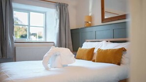 A neatly made bed with white and yellow pillows, a window with curtains, and a lamp on a dresser. - Cosy Country Cottage Sleeps 4 (Kirkbymoorside)