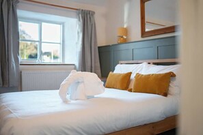 A neatly made bed with white and yellow pillows, a window with curtains, and a lamp on a dresser. - Cosy Country Cottage Sleeps 4 (Kirkbymoorside)