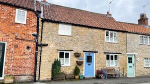A two-story stone building with a red-tiled roof, a blue door, and a small garden area with potted plants. - Cosy Country Cottage Sleeps 4 (Kirkbymoorside)