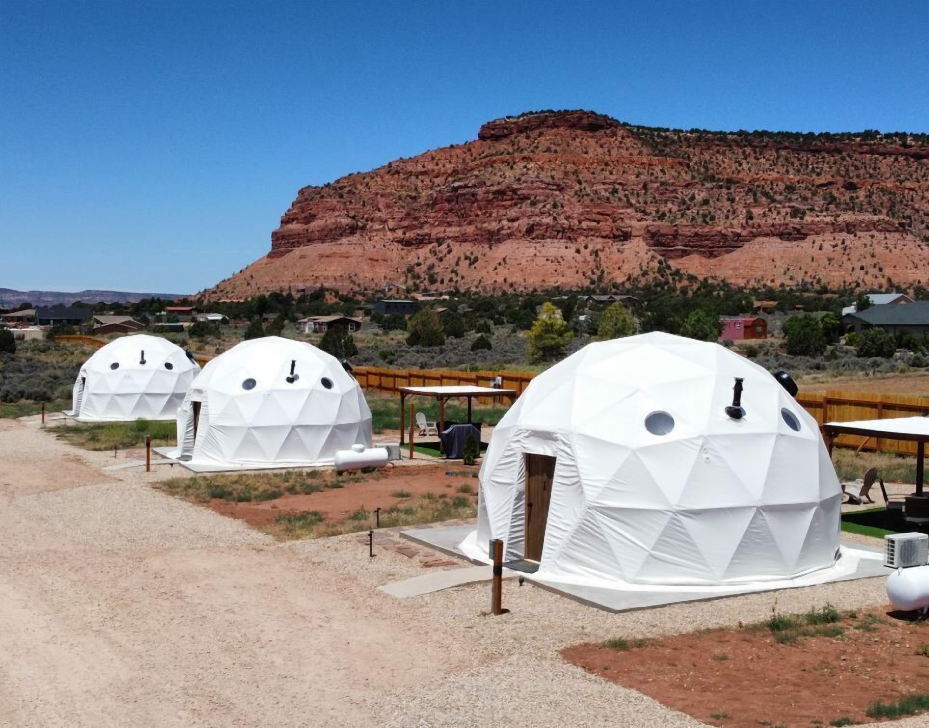 Three dome-shaped structures with a geometric pattern, set against a backdrop of a red rock formation and a clear blue sky.