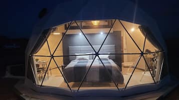 A geodesic dome structure with a bedroom featuring a bed, bedside table, and a chair.