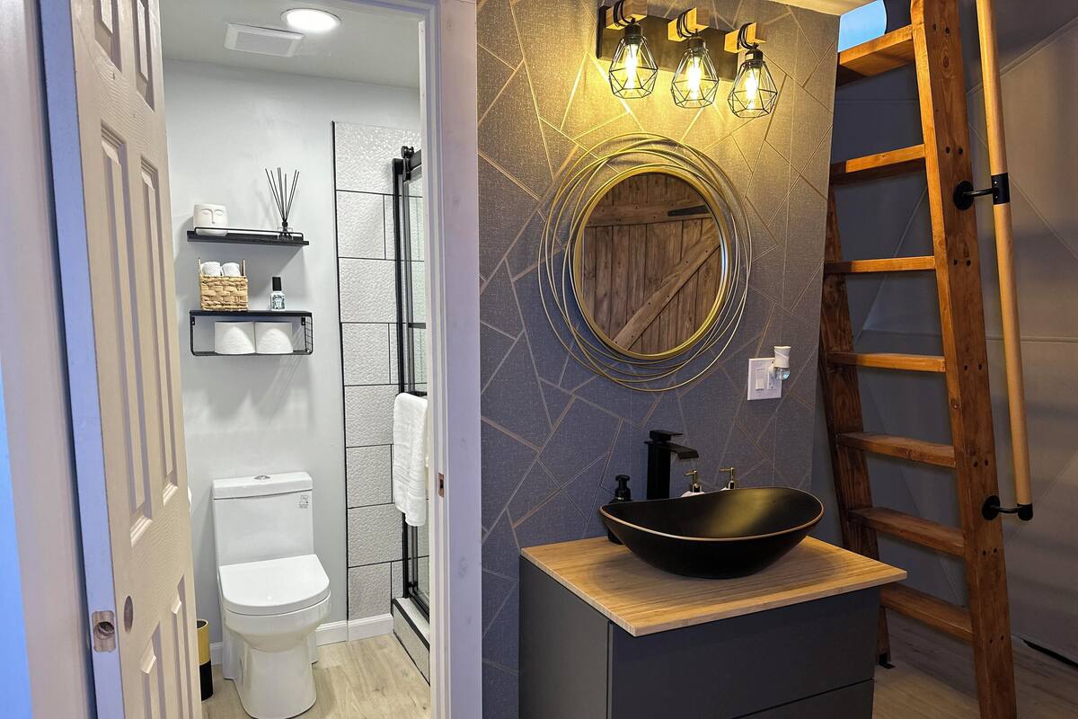 A bathroom with a white toilet, a wooden vanity with a black sink, a wooden ladder, and a wall-mounted mirror with geometric lighting.