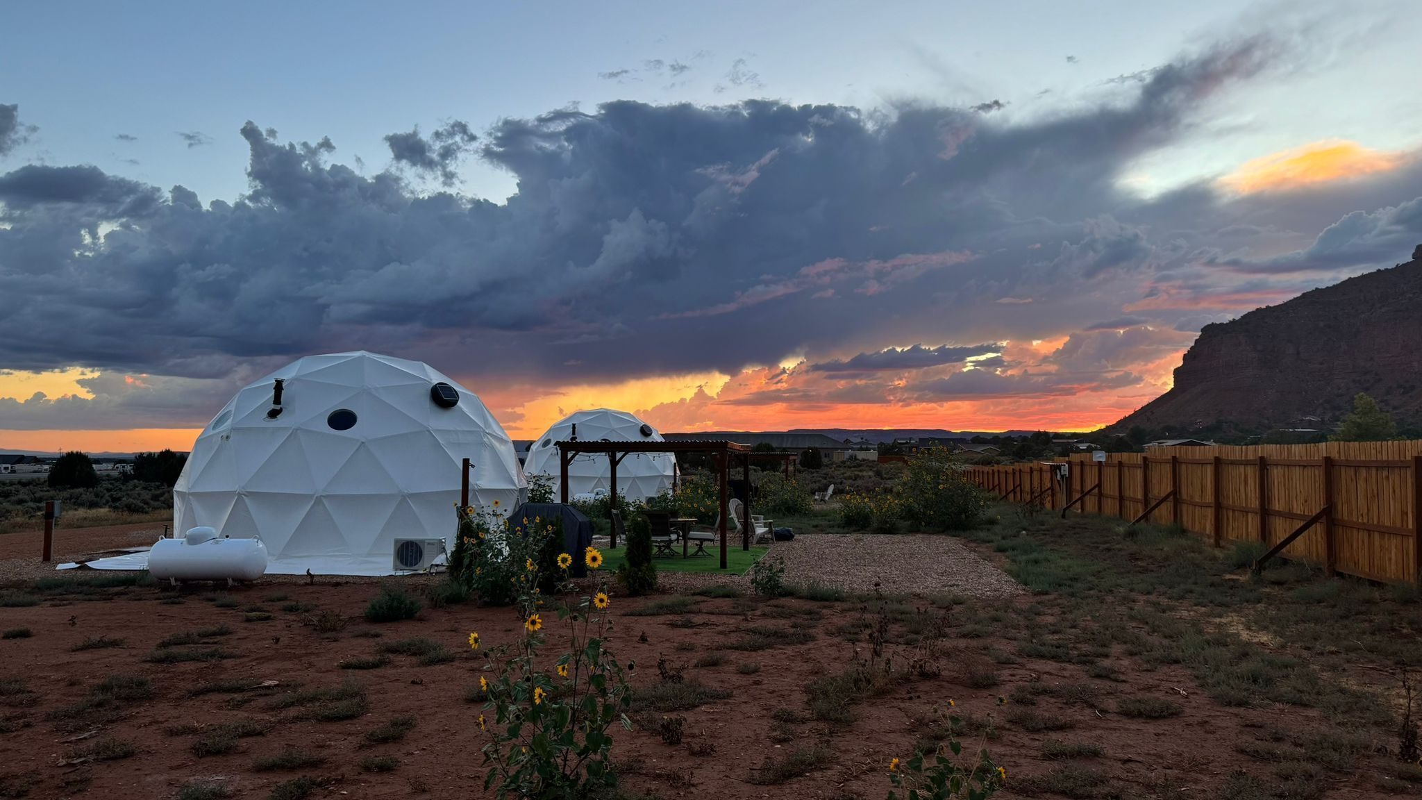 Dos estructuras de domo geodésico con un pérgola de madera y una valla de madera en un paisaje desértico al atardecer.
