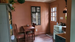 Een eethoek met een houten tafel en stoelen, een raam met luiken, een kitchenette met een koelkast, en een hangplant.