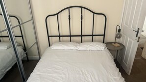 Uma cama de solteiro com estrutura de metal e roupa de cama branca num quarto com porta branca e uma pequena mesa de cabeceira com uma lâmpada.